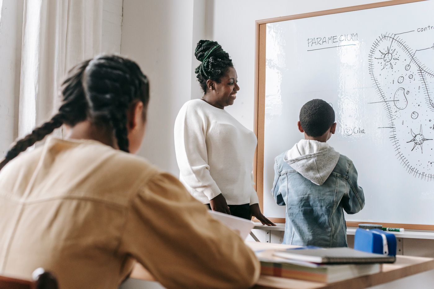 Une enseignante accompagne un élève au tableau pendant qu’une camarade suit la leçon